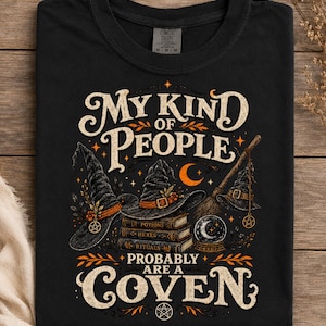 May include: A black t-shirt featuring a graphic print with the text "MY KIND OF PEOPLE PROBABLY ARE A COVEN". The design includes three witch hats, a stack of books labeled "POTIONS", "HEXES", and "RITUALS", a broom, a crystal ball, and a crescent moon.