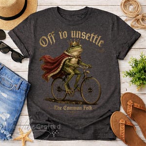 Puede incluir: Camiseta gris oscuro con una ilustración caprichosa de una rana con corona y capa, montando en bicicleta. El texto "Off to unsettle - The Common Folk" está arqueado. Una prenda informal, adecuada para el uso diario.