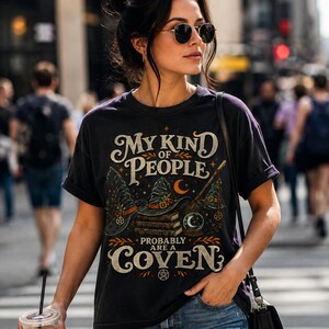 May include: A black short-sleeve t-shirt featuring a graphic print with the text "MY KIND OF PEOPLE PROBABLY ARE A COVEN". The design includes illustrations of witch hats, a stack of books, a broomstick, a crystal ball, and small pentagrams.