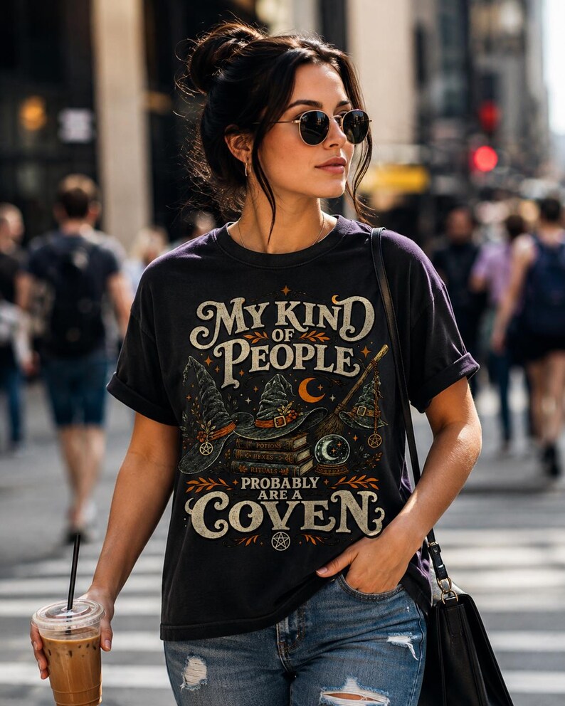 May include: A black short-sleeve t-shirt featuring a graphic print with the text "MY KIND OF PEOPLE PROBABLY ARE A COVEN". The design includes illustrations of witch hats, a stack of books, a broomstick, a crystal ball, and small pentagrams.