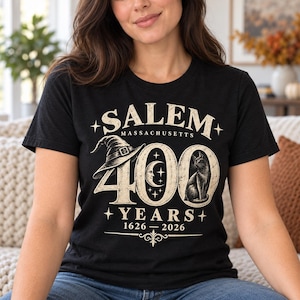 May include: A black short-sleeved t-shirt featuring a cream-colored graphic that reads SALEM MASSACHUSETTS 400 YEARS 1626-2026. The design includes a witch hat, a crescent moon, stars, and a cat illustration integrated into the large number 400.