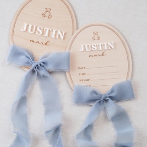May include: Two oval wooden plaques featuring a light wood grain, each adorned with a dusty blue frayed ribbon bow. The plaques display a small bear icon, the name JUSTIN mark in white and brown text, and lines for date, time, weight, and length.