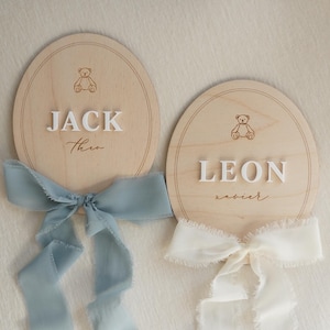 May include: Two oval wooden plaques featuring engraved teddy bear icons and raised white lettering. One plaque displays the names JACK and Theo with a dusty blue frayed ribbon, while the other shows LEON and xavier with a cream frayed ribbon.