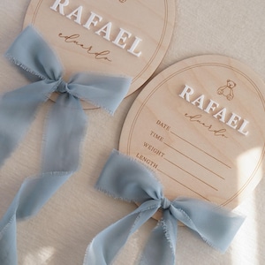 May include: Two arched wooden plaques featuring a laser-engraved teddy bear icon and the name RAFAEL eduardo in white 3D lettering. Each plaque is adorned with a dusty blue frayed chiffon ribbon. One plaque includes blank lines for date, time, weight, and length.