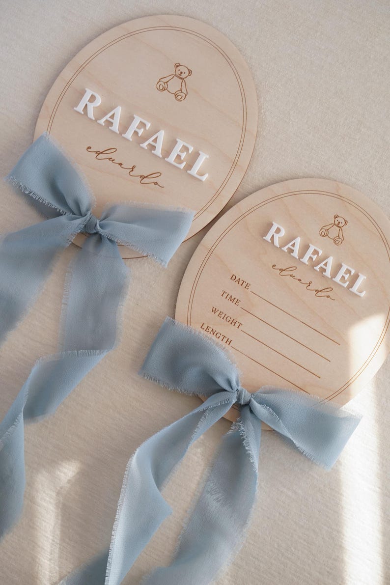 May include: Two arched wooden plaques featuring a laser-engraved teddy bear icon and the name RAFAEL eduardo in white 3D lettering. Each plaque is adorned with a dusty blue frayed chiffon ribbon. One plaque includes blank lines for date, time, weight, and length.