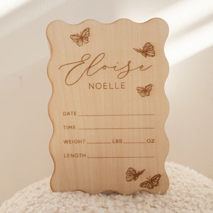 May include: A light-colored wooden sign with a wavy edge, featuring the name "Eloise Noelle" and butterfly illustrations. The sign includes spaces to record birth details: date, time, weight (lbs, oz), and length.