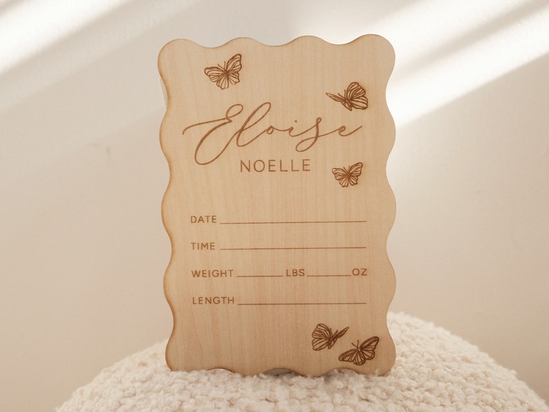 May include: A light-colored wooden sign with a wavy edge, featuring the name "Eloise Noelle" and butterfly illustrations. The sign includes spaces to record birth details: date, time, weight (lbs, oz), and length.