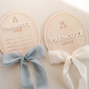 May include: Two oval wooden birth announcement plaques featuring a laser-engraved teddy bear icon and the name PRESCOTT LOUIS in raised white lettering. The left plaque includes lines for date, time, weight, and length, adorned with a dusty blue ribbon.