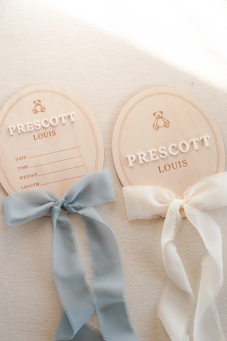 May include: Two oval wooden birth announcement plaques featuring a laser-engraved teddy bear icon and the name PRESCOTT LOUIS in raised white lettering. The left plaque includes lines for date, time, weight, and length, adorned with a dusty blue ribbon.
