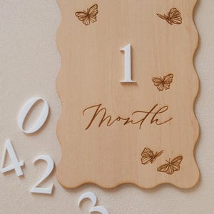 May include: A wooden milestone plaque with a wavy edge, featuring the number "1" and the word "Month" in script. The plaque is decorated with etched butterfly designs. White numbers 0, 2, 3, 4, and 5 are scattered around the plaque.