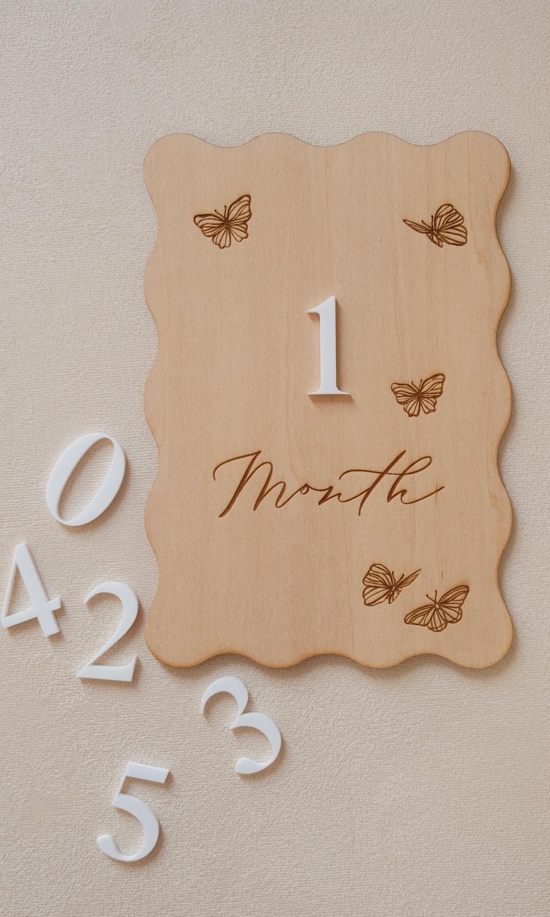 May include: A wooden milestone plaque with a wavy edge, featuring the number "1" and the word "Month" in script. The plaque is decorated with etched butterfly designs. White numbers 0, 2, 3, 4, and 5 are scattered around the plaque.