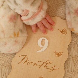 May include: A wooden milestone plaque with the number "9" and the word "Months" in script. The plaque has a wavy edge and butterfly illustrations. A baby's hand rests on the plaque, with a soft, floral-patterned blanket in the background.