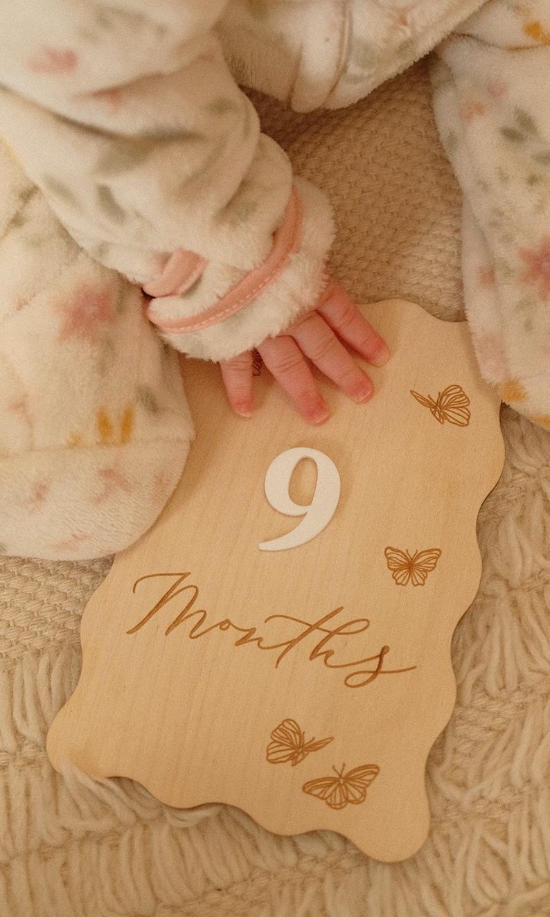 May include: A wooden milestone plaque with the number "9" and the word "Months" in script. The plaque has a wavy edge and butterfly illustrations. A baby's hand rests on the plaque, with a soft, floral-patterned blanket in the background.