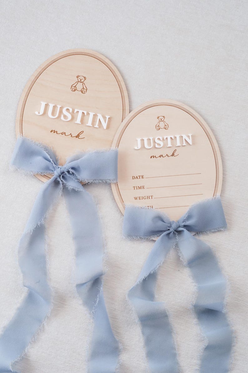 May include: Two oval wooden plaques featuring a light wood grain, each adorned with a dusty blue frayed ribbon bow. The plaques display a small bear icon, the name JUSTIN mark in white and brown text, and lines for date, time, weight, and length.