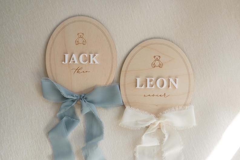 May include: Two oval wooden plaques featuring engraved teddy bear icons and raised white lettering. One plaque displays the names JACK and Theo with a dusty blue frayed ribbon, while the other shows LEON and xavier with a cream frayed ribbon.