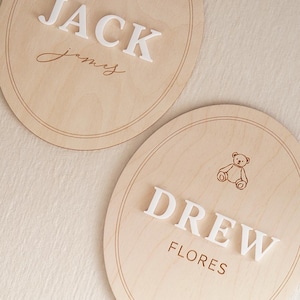 May include: Two oval light wood plaques featuring engraved teddy bear illustrations and double-line borders. One plaque displays the white 3D text JACK and the script name James. The other plaque displays the white 3D text DREW and the name FLORES.