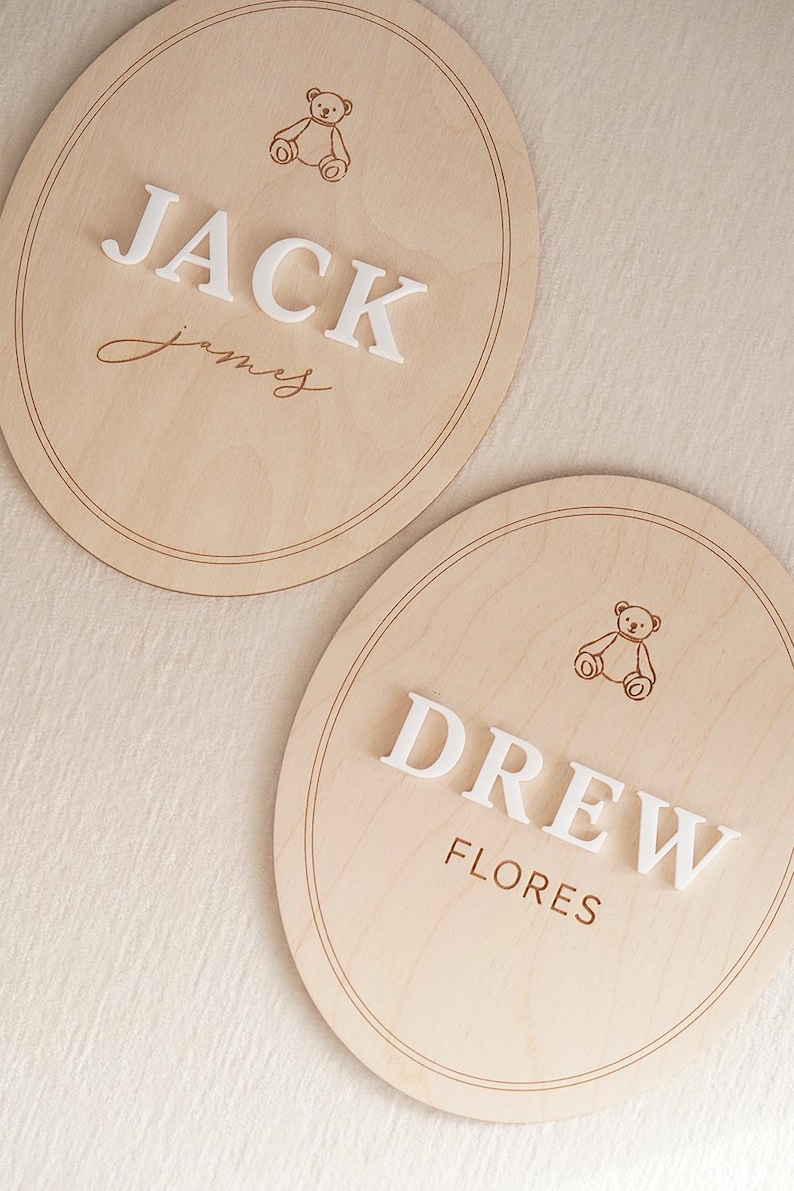 May include: Two oval light wood plaques featuring engraved teddy bear illustrations and double-line borders. One plaque displays the white 3D text JACK and the script name James. The other plaque displays the white 3D text DREW and the name FLORES.