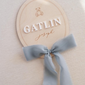 May include: An oval wooden plaque featuring a laser-engraved teddy bear icon, the white 3D-style text GATLIN, and the script text joseph in brown. A dusty blue frayed-edge ribbon is tied in a bow at the bottom of the light-coloured wooden sign.