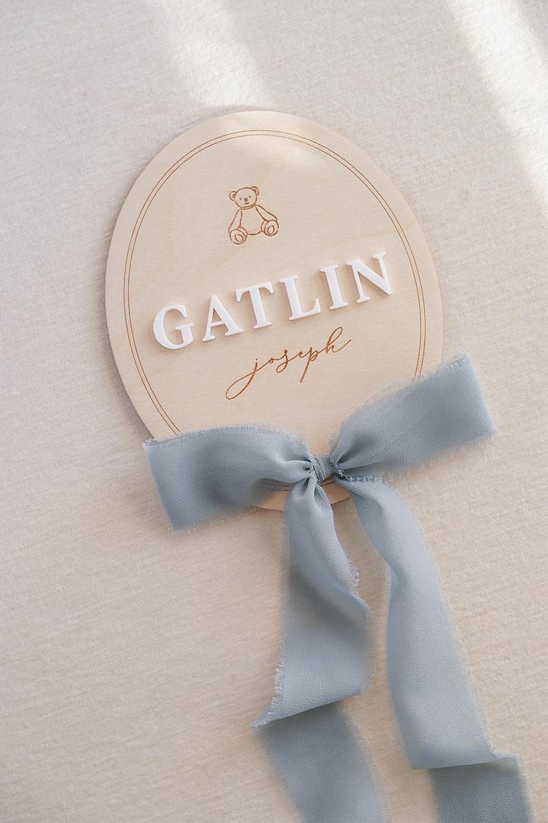 May include: An oval wooden plaque featuring a laser-engraved teddy bear icon, the white 3D-style text GATLIN, and the script text joseph in brown. A dusty blue frayed-edge ribbon is tied in a bow at the bottom of the light-coloured wooden sign.