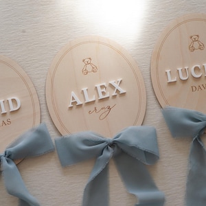 May include: Three oval light-coloured wooden plaques featuring engraved teddy bear icons and raised white lettering. The plaques display the names DAVID THOMAS, ALEX cruz, and LUCIAN DAVID. Each plaque is adorned with a dusty blue chiffon ribbon bow.
