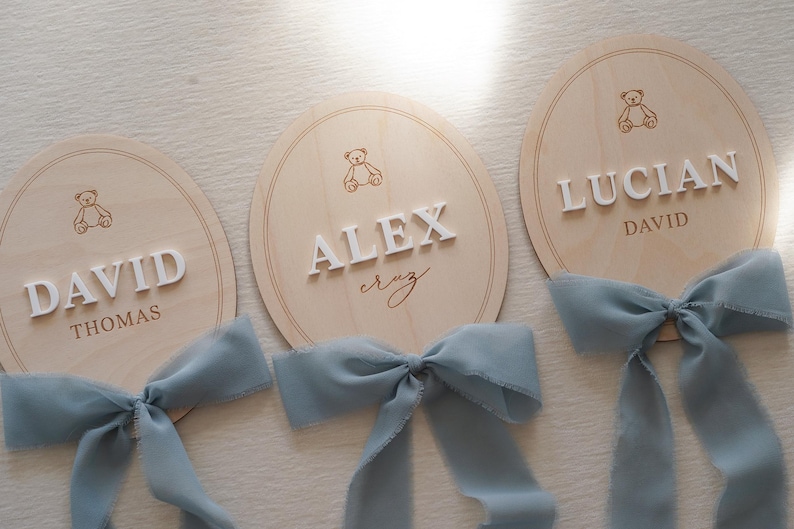 May include: Three oval light-coloured wooden plaques featuring engraved teddy bear icons and raised white lettering. The plaques display the names DAVID THOMAS, ALEX cruz, and LUCIAN DAVID. Each plaque is adorned with a dusty blue chiffon ribbon bow.