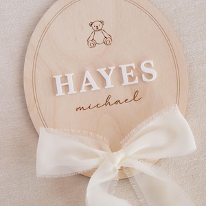 May include: A circular light-coloured wooden plaque featuring an engraved teddy bear icon at the top, the white raised 3D lettering HAYES in the centre, the name michael in cursive script below, and a sheer white ribbon tied at the bottom edge.