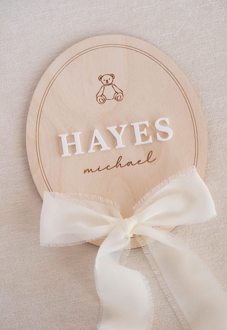 May include: A circular light-coloured wooden plaque featuring an engraved teddy bear icon at the top, the white raised 3D lettering HAYES in the centre, the name michael in cursive script below, and a sheer white ribbon tied at the bottom edge.