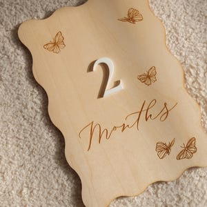 May include: Wooden milestone plaque with a wavy edge, featuring the number "2" in white and the word "Months" in script. Delicate butterfly illustrations are etched around the edges, perfect for baby photos. The plaque is made of light-colored wood.