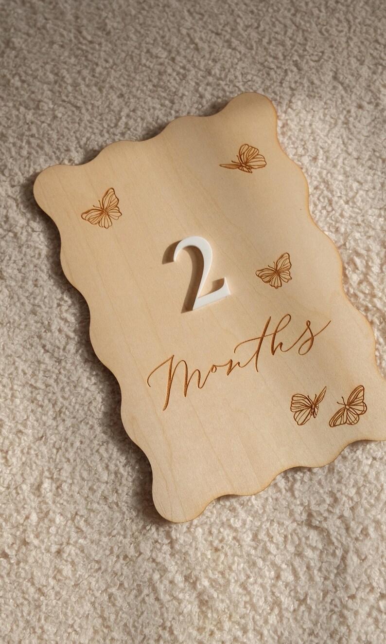 May include: Wooden milestone plaque with a wavy edge, featuring the number "2" in white and the word "Months" in script. Delicate butterfly illustrations are etched around the edges, perfect for baby photos. The plaque is made of light-colored wood.