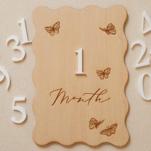May include: A wooden milestone plaque with a wavy edge, featuring the number "1" and the word "Month" in script. Delicate butterfly illustrations are etched on the light-colored wood. White numbers 2, 3, 5 are visible.