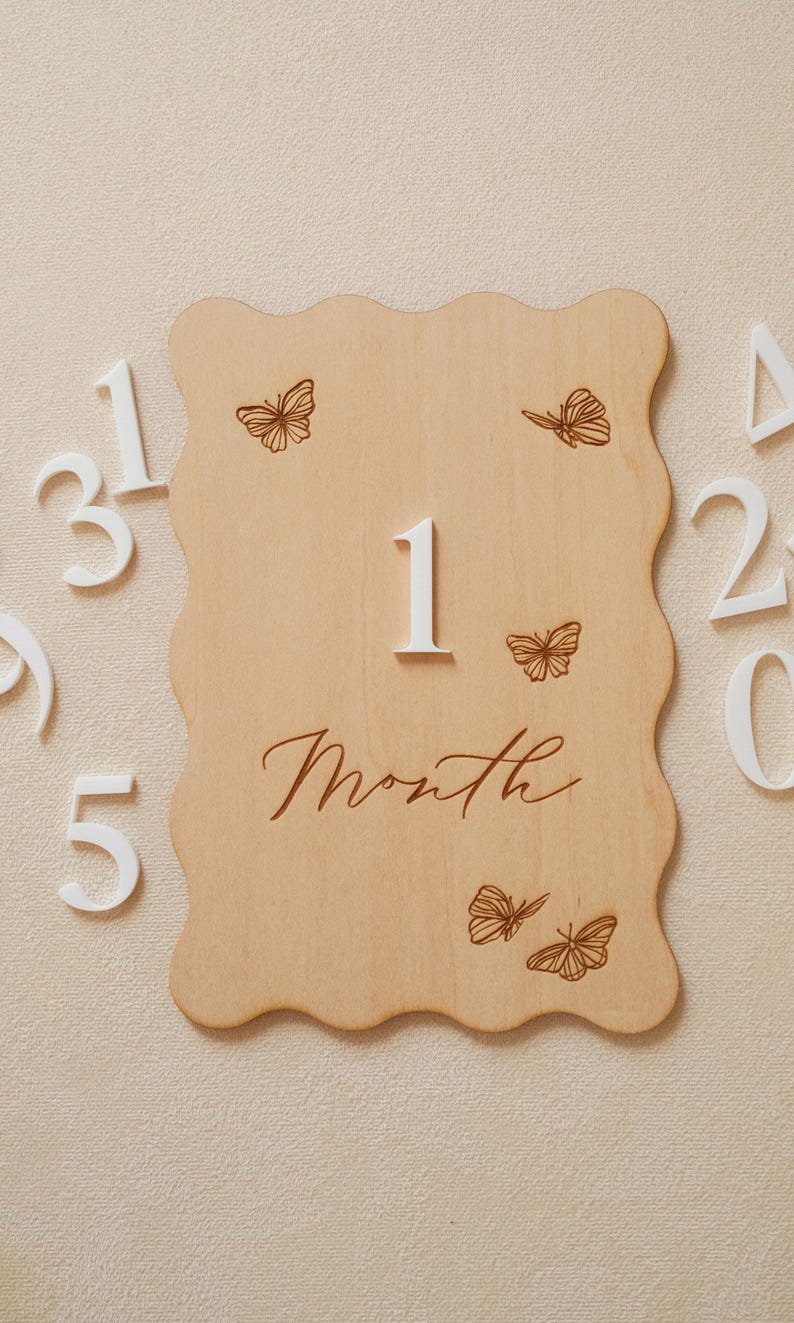 May include: A wooden milestone plaque with a wavy edge, featuring the number "1" and the word "Month" in script. Delicate butterfly illustrations are etched on the light-colored wood. White numbers 2, 3, 5 are visible.