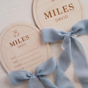 May include: Two round light-coloured wooden plaques featuring an engraved teddy bear icon and the name MILES DAVID. One plaque includes blank lines for DATE, TIME, WEIGHT, and LENGTH. Each plaque is adorned with a dusty blue frayed silk ribbon.
