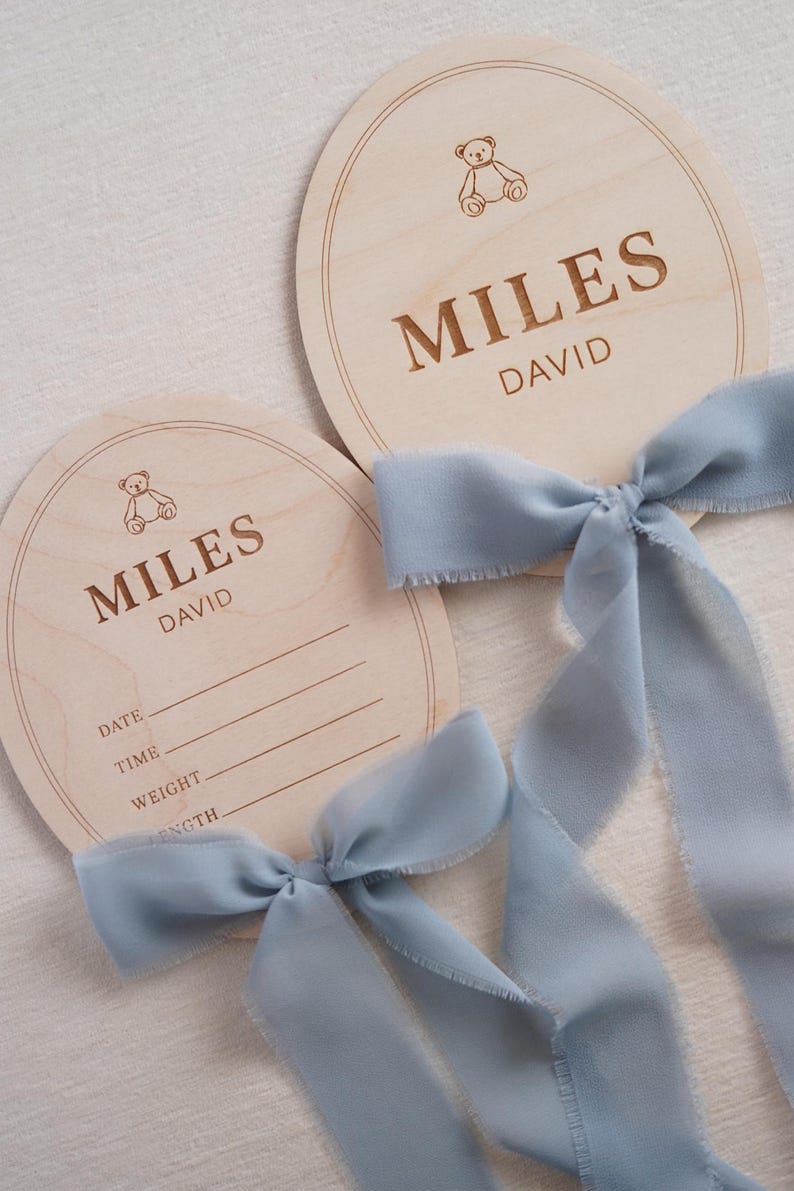 May include: Two round light-coloured wooden plaques featuring an engraved teddy bear icon and the name MILES DAVID. One plaque includes blank lines for DATE, TIME, WEIGHT, and LENGTH. Each plaque is adorned with a dusty blue frayed silk ribbon.