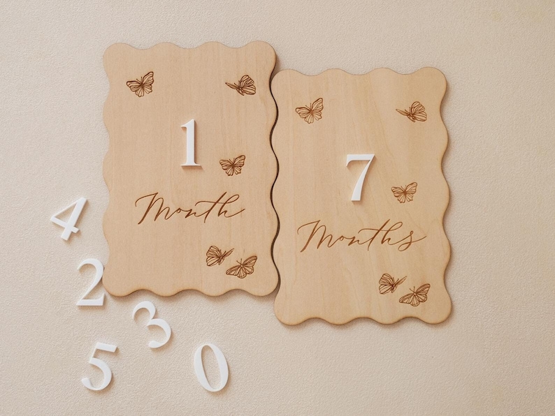 May include: Two wooden milestone plaques with scalloped edges, featuring engraved butterflies and the numbers "1" and "7" with the words "Month" and "Months". Additional white numbers are scattered around. The plaques are light brown.