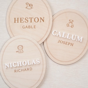 May include: Three oval light-coloured wooden plaques featuring engraved borders and icons. One plaque displays HESTON GABLE with a rocking horse. Another shows CALLUM JOSEPH with a teddy bear. The third features NICHOLAS RICHARD with a lion icon.