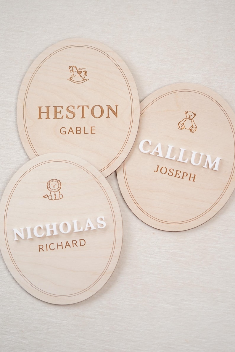 May include: Three oval light-coloured wooden plaques featuring engraved borders and icons. One plaque displays HESTON GABLE with a rocking horse. Another shows CALLUM JOSEPH with a teddy bear. The third features NICHOLAS RICHARD with a lion icon.