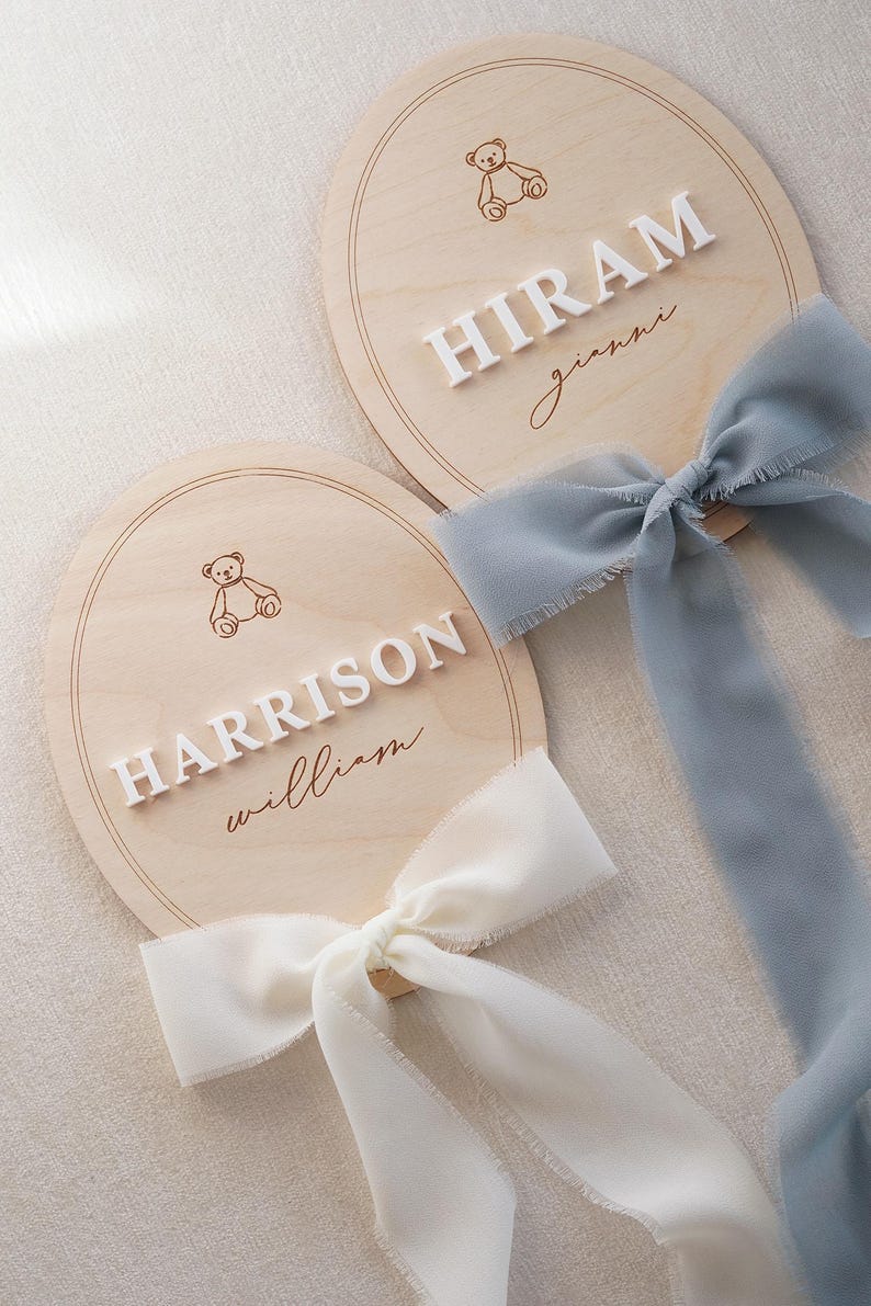 May include: Two round wooden plaques featuring engraved teddy bear illustrations and raised white lettering. One plaque displays the names HIRAM and gianni with a dusty blue ribbon, while the other shows HARRISON and william with a cream ribbon.