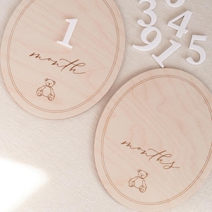May include: Two oval light wood milestone plaques featuring a thin engraved border, a line drawing of a teddy bear, and the script text month or months. Includes a set of separate white wooden numbers 1, 3, 4, 5, 6, 7, and 9 for customisation.