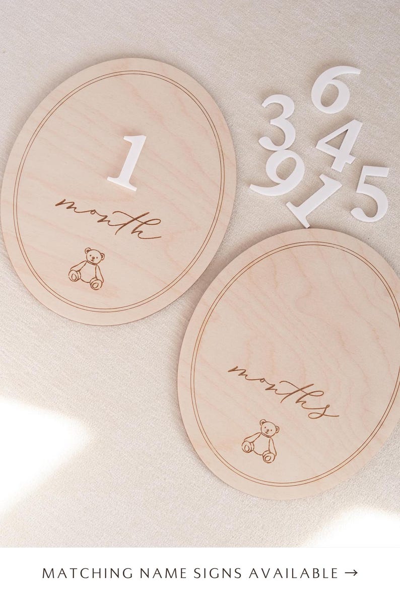 May include: Two oval light wood milestone plaques featuring a thin engraved border, a line drawing of a teddy bear, and the script text month or months. Includes a set of separate white wooden numbers 1, 3, 4, 5, 6, 7, and 9 for customisation.