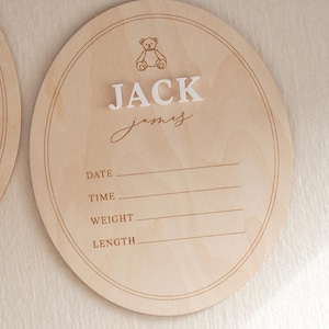 May include: A round light-coloured wooden birth announcement sign featuring an engraved teddy bear icon, the name JACK JAMES in raised white lettering, and printed lines for date, time, weight, and length with double-line circular borders.