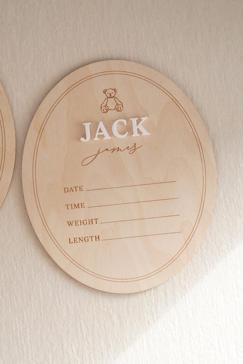 May include: A round light-coloured wooden birth announcement sign featuring an engraved teddy bear icon, the name JACK JAMES in raised white lettering, and printed lines for date, time, weight, and length with double-line circular borders.