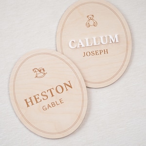 May include: Two oval light-coloured wooden plaques featuring engraved borders. One plaque displays a brown engraved teddy bear icon above the white 3D text CALLUM and brown text JOSEPH. The other shows a brown rocking horse icon above HESTON GABLE.