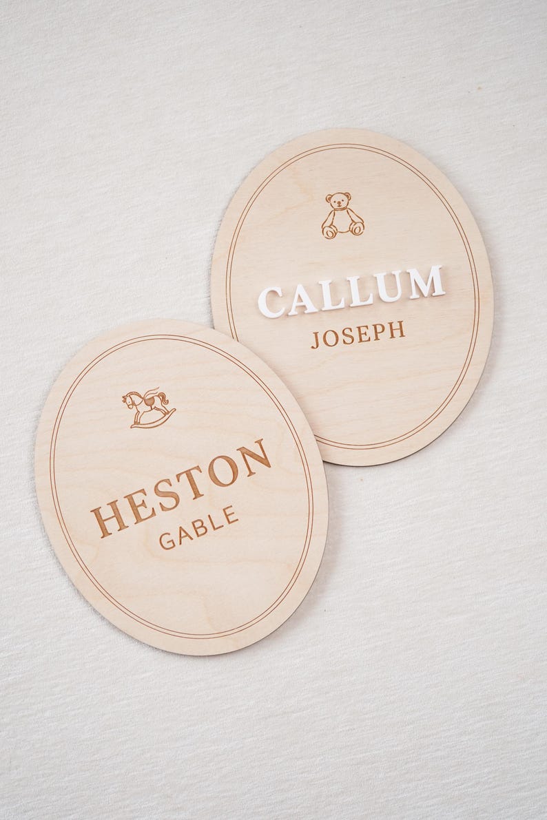 May include: Two oval light-coloured wooden plaques featuring engraved borders. One plaque displays a brown engraved teddy bear icon above the white 3D text CALLUM and brown text JOSEPH. The other shows a brown rocking horse icon above HESTON GABLE.