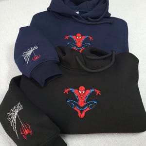 May include: A navy blue hoodie and a black hoodie, each featuring an embroidered red and blue superhero figure on the chest. The sleeves of both hoodies display an embroidered white spiderweb design with a small red spider hanging from the web.