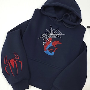 Może przedstawiać: Granatowa bluza z kapturem z haftowanym wzorem Spider-Mana. Z przodu Spider-Man z pajęczyną, a na lewym rękawie czerwony emblemat pająka. Wygodny, codzienny element garderoby.