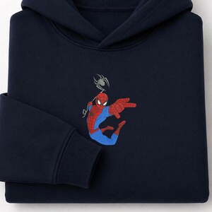 May include: A dark navy blue hooded sweatshirt featuring an embroidered graphic of a red and blue superhero character in a dynamic pose with a spider web line extending upward to a small grey spider positioned above the character's head.