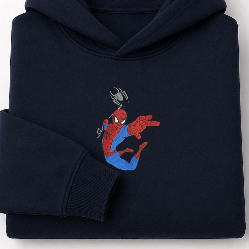 May include: A dark navy blue hooded sweatshirt featuring an embroidered graphic of a red and blue superhero character in a dynamic pose with a spider web line extending upward to a small grey spider positioned above the character's head.