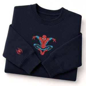 May include: Navy blue sweatshirt featuring a Spider-Man graphic in red, blue, and black. The superhero is depicted in a dynamic pose. A red spider emblem is on the left sleeve. This crewneck sweatshirt is a great gift for fans of the Marvel comic book character.