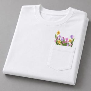 K&ouml;nnte beinhalten: Wei&szlig;es Sweatshirt mit Rundhalsausschnitt und einer Tasche mit farbenfroher Blumenstickerei. Die Stickerei umfasst gelbe, rosa und lila Blumen mit gr&uuml;nen Stielen. Das Sweatshirt ist sauber auf einer grauen Oberfl&auml;che gefaltet.
