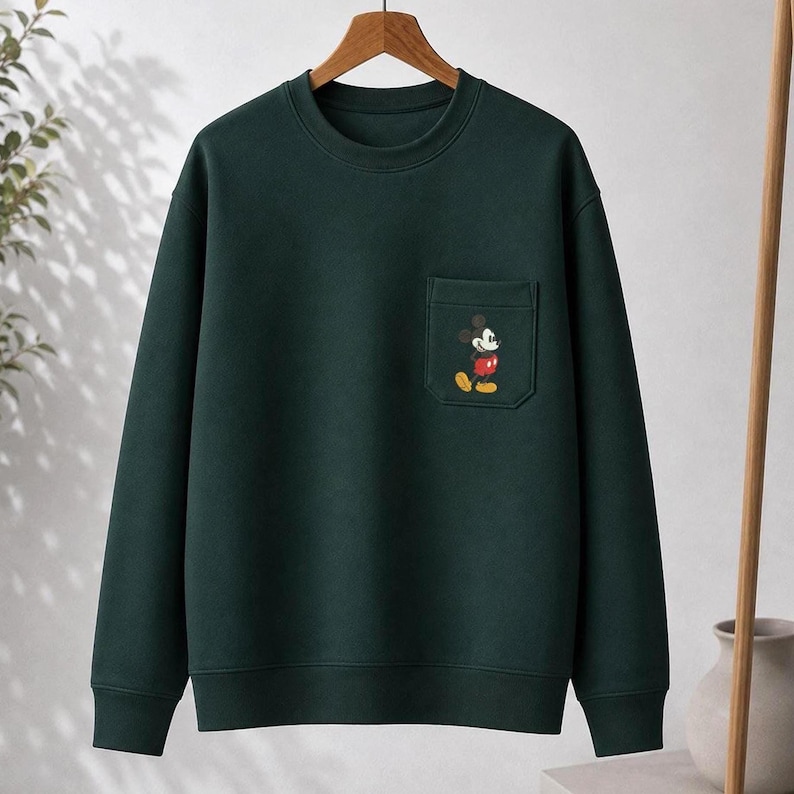 May include: A dark forest green crewneck sweatshirt featuring a single chest pocket on the left side. The pocket is adorned with a small, embroidered graphic of Mickey Mouse wearing red shorts and yellow shoes, set against the solid green fabric.
